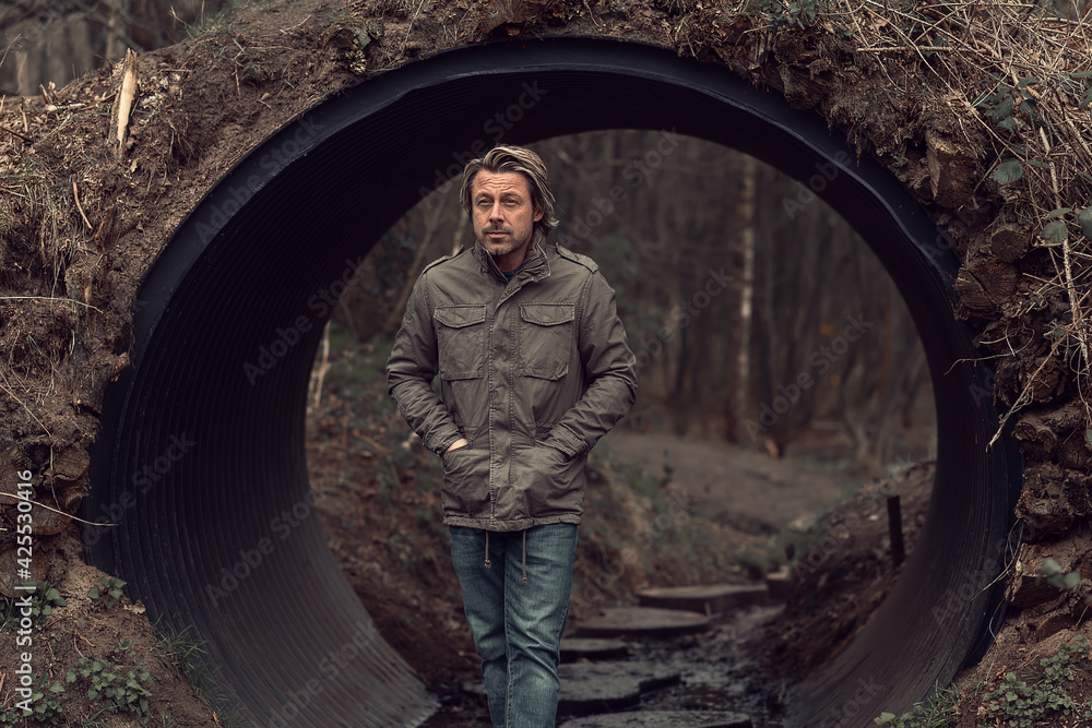 Blond man in green jacket and jeans stands in a tube tunnel in nature reserve. Blond man in green jacket and jeans stands in a tube tunnel in nature reserve.