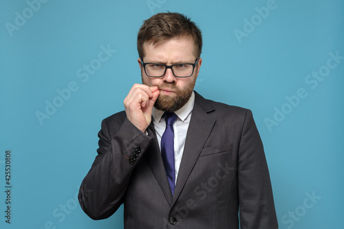Suspicious Caucasian man in business suit, sternly and suspiciously looks, touching mustache with hands. Blue background.