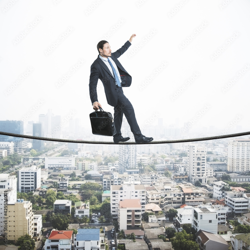 Business risk concept with businessman in black suit with briefcase walking the tightrope over cityscape Business risk concept with businessman in black suit with briefcase walking the tightrope over cityscape