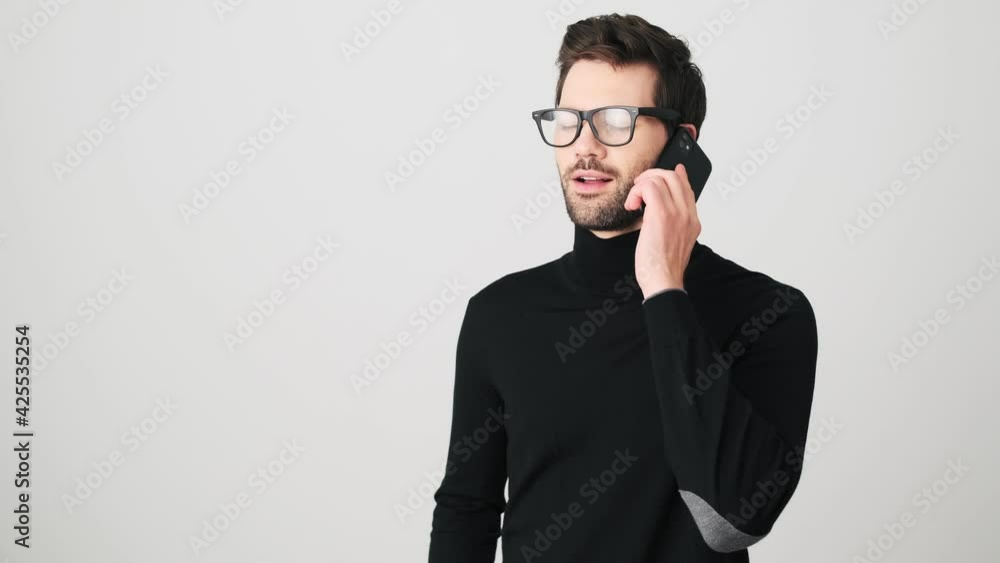 A surprised man is talking on his smartphone while doing winner gesture standing isolated over a white wall in a studio A surprised man is talking on his smartphone while doing winner gesture standing isolated over a white wall in a studio