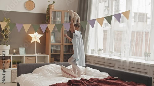 Full shot of petite adorable Mixed-Race girl wearing hair in bun, holding toy bear, jumping on bed in her room
