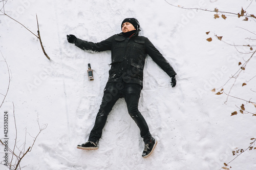 A severely drunk homeless man in a black coat and hat lies, asleep on his back on white snow in the cold winter with a bottle of alcohol. Photography, concept.