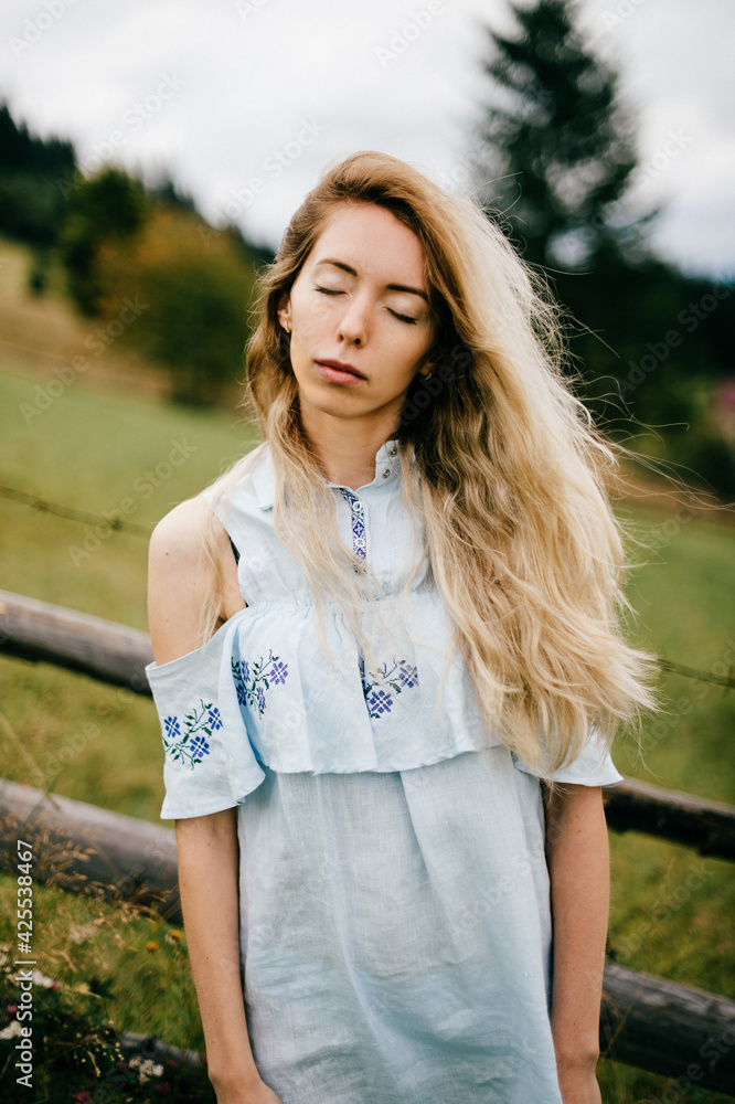 Young attractive elegant blonde girl in blue romantic dress posing near fence in the countryside Young attractive elegant blonde girl in blue romantic dress posing near fence in the countryside