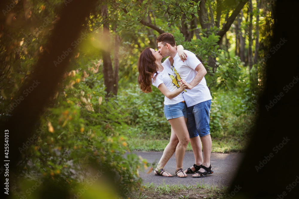 A young couple in love walks in a summer park at sunset and kisses and hugs hard. The heterosexual couple is wearing the same outfit, white T-shirts and shorts. Tender relationship between lovers A young couple in love walks in a summer park at sunset and kisses and hugs hard. The heterosexual couple is wearing the same outfit, white T-shirts and shorts. Tender relationship between lovers