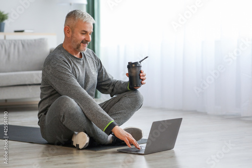 Senior man in sportswear sitting on fitness mat, using laptop