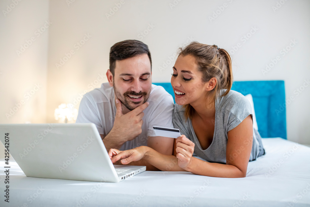 Fototapeta premium Young couple is browsing the internet on a laptop. Couple is shopping online using laptop and credit card in the bed. Beautiful young amorous couple using laptop, on bed.