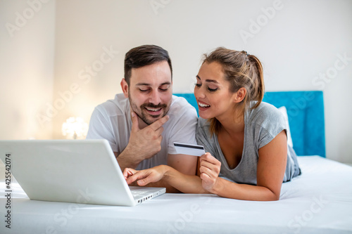 Young couple is browsing the internet on a laptop. Couple is shopping online using laptop and credit card in the bed. Beautiful young amorous couple using laptop, on bed.