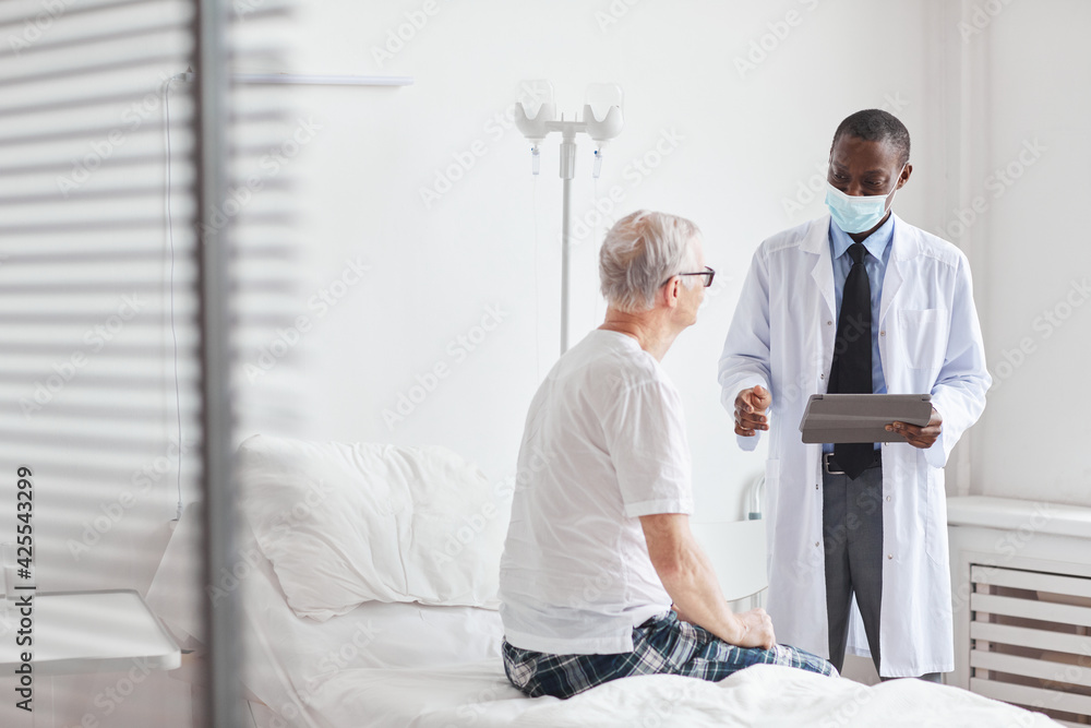 Portrait of African-American doctor talking to senior man in white hospital room, copy space
