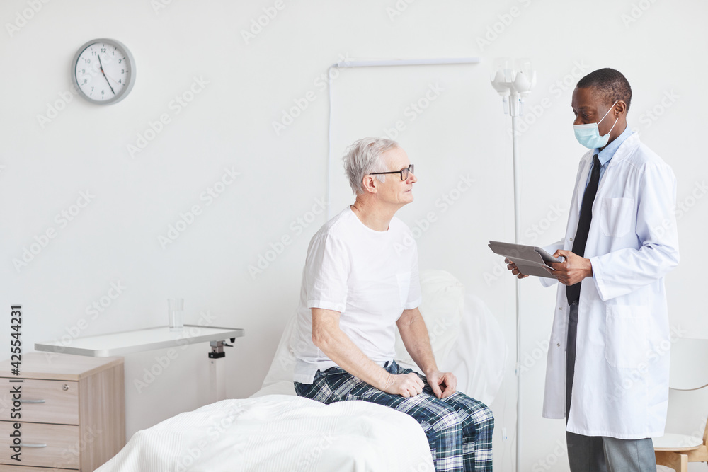 Obraz premium Portrait of recovering senior man talking to African-American doctor while sitting on bed in white hospital room, copy space