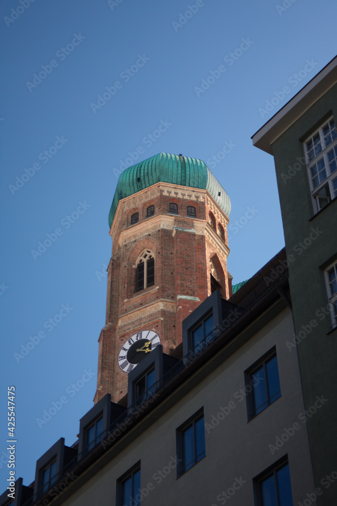 München: Der Turm der Frauenkirche an einem sonnigen Tag mit blauem Himmel. Kontrast zwischen den gewöhnlichen Häusern im Vordergrund und der prächtigen Kirche im Hintergrund 