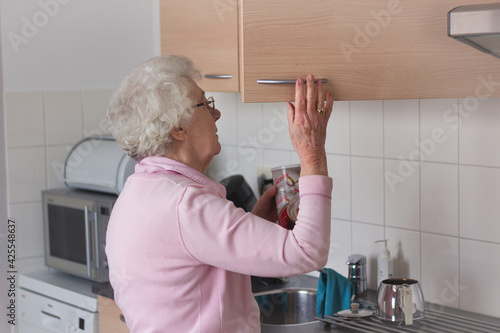 An old senior woman, 80s adult, grandmother, standing in a kitchen, opening a kitchen cabinet door and making coffee and tea. Retirement, aging and self-sustainability concept.