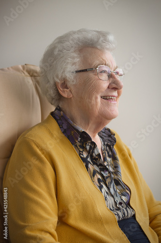 Vertical portrait of an old senior woman, 80s adult, grandmother, sitting in a chair on her balcony, smiling, thinking and looking in the distance.
