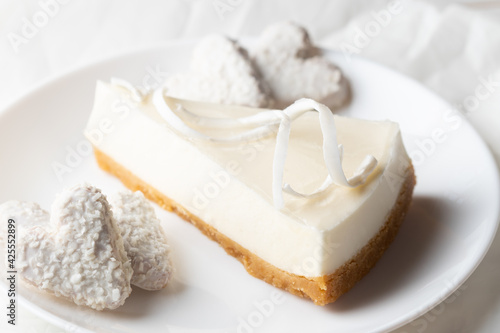 Vegan cheesecake with coconut. A piece of cheesecake is on a white plate. Nearby are vegan heart-shaped cookies. Close-up. Photography in white colors