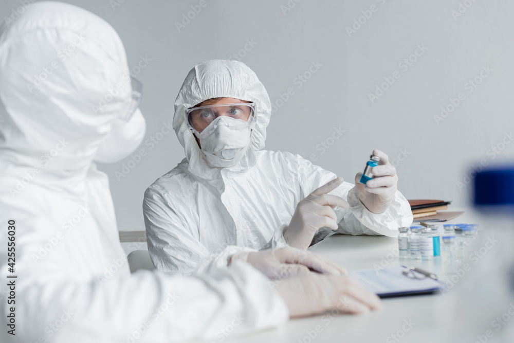Scientist in protective suit pointing at vaccine near colleague on blurred foreground in laboratory Scientist in protective suit pointing at vaccine near colleague on blurred foreground in laboratory