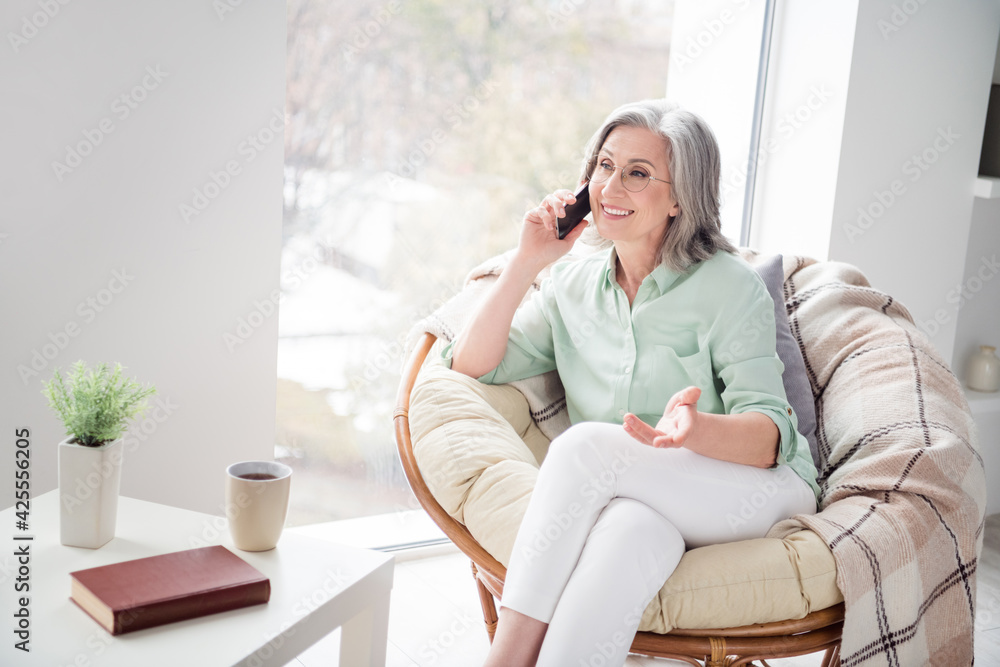 Photo of pretty positive person sit on chair near window speak phone have good mood weekend indoors Photo of pretty positive person sit on chair near window speak phone have good mood weekend indoors