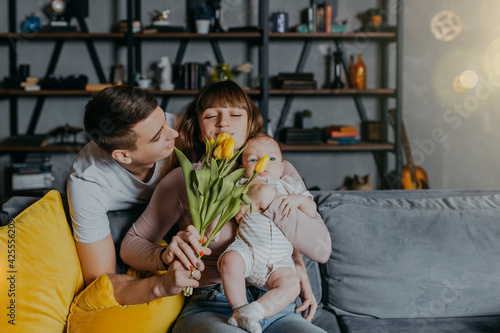 Man gives flowers to woman with little kid in arms. Mother's Day congratulations. Happy family with flowers on couch. Bunch of yellow tulips.
