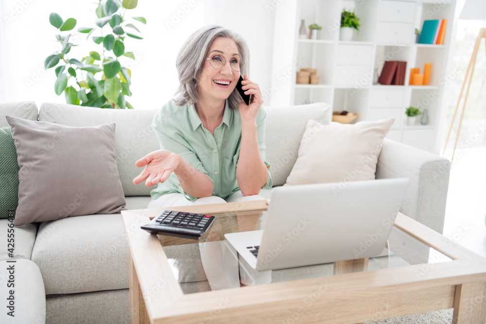 Photo of cheerful person sit on couch speak phone arm palm explain tell have good mood toothy smile indoors Photo of cheerful person sit on couch speak phone arm palm explain tell have good mood toothy smile indoors