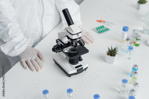 Cropped view of scientist in hazmat suit standing near microscope and vaccines on table