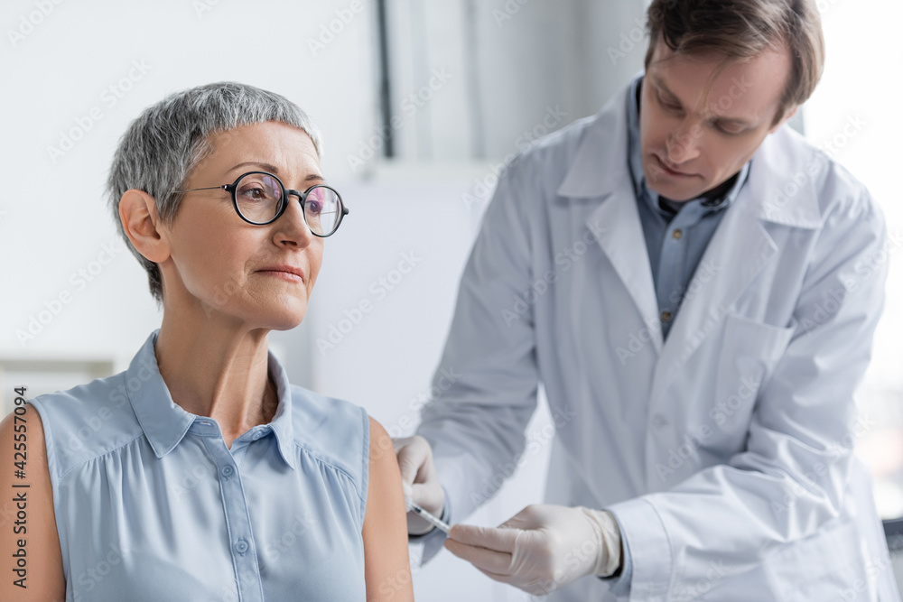 Mature patient in eyeglasses looking away near doctor doing vaccination on blurred background Mature patient in eyeglasses looking away near doctor doing vaccination on blurred background