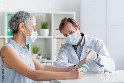 Doctor in medical mask pointing at pills near mature patient and vaccine on blurred foreground