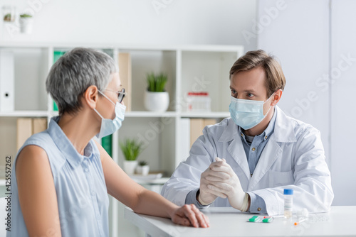 Doctor in medical mask sitting near mature patient and vaccine with pills during consultation