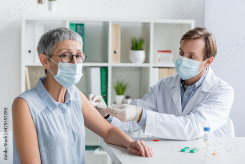 Doctor in medical mask doing vaccination of mature patient on blurred foreground near pills on table