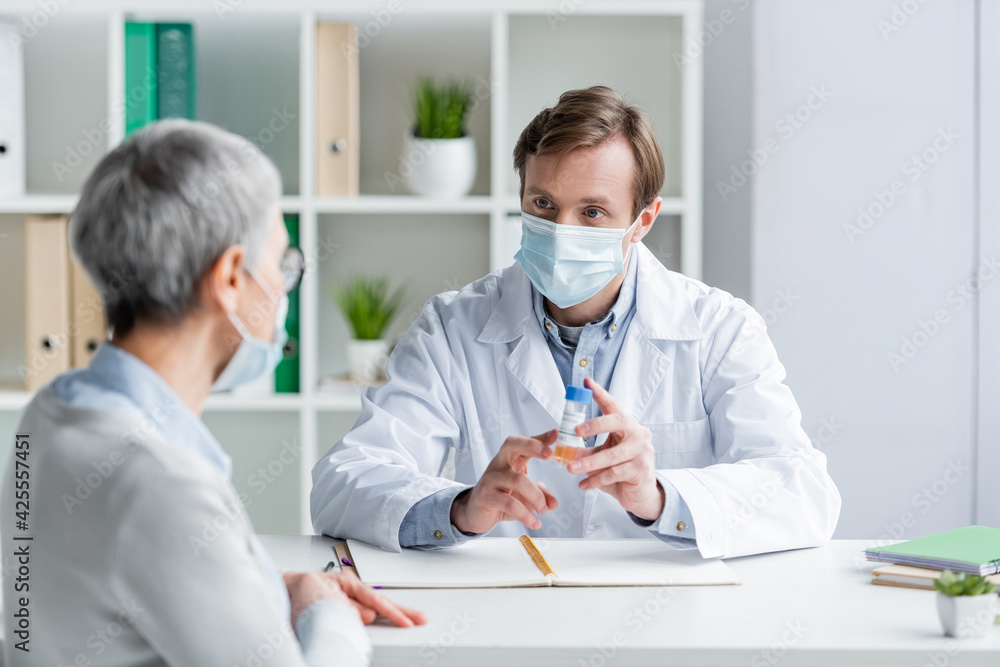 © LIGHTFIELD STUDIOS - Doctor in medical mask holding vaccine near mature patient on blurred foreground © LIGHTFIELD STUDIOS - Doctor in medical mask holding vaccine near mature patient on blurred foreground