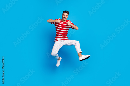 Full body photo of happy brunette positive young man show thumbs-up jump air isolated on blue color background