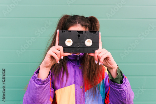 Young girl in a sports jacket from the 90s. Fashionable woman near a green wall holds a videotape in crayfish. Fashion trends, street look. VHS Videotapes. Let's go back to the 90s 80s