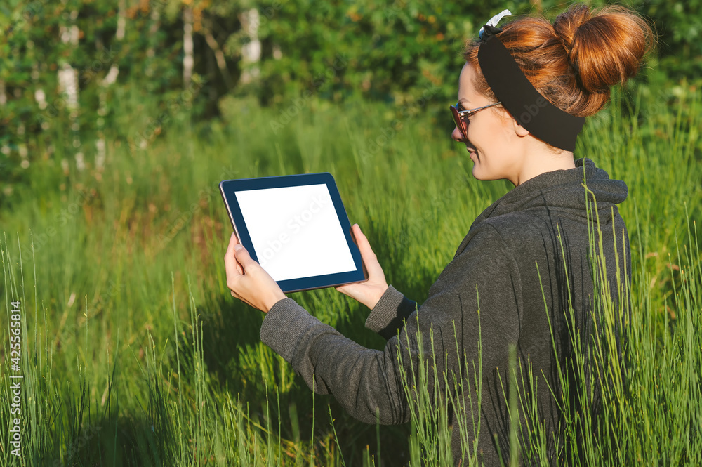 Happy girl holding a mock up tablet in a meadow near the forest.