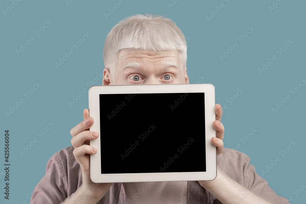 Albino guy hiding behind tablet computer with blank screen on turquoise studio background, mockup for design