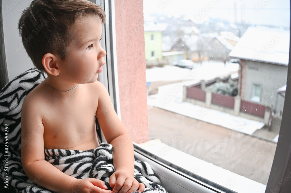 Beautiful little boy sits on the windowsill and looks out the window. Beautiful little boy sits on the windowsill and looks out the window.