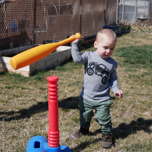 A toddler boy watches his baseball fly away after knocking it off the tee with a plastic bat on a sunny spring day. 
