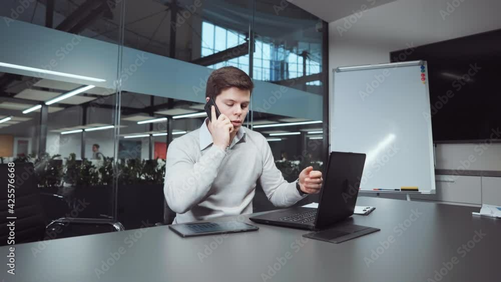 Young man wearing sweater working in office, using laptop, talking on phone and swiping screens on tablet, unrecognizable people walking behind glass wall. Financial consultant at workplace Young man wearing sweater working in office, using laptop, talking on phone and swiping screens on tablet, unrecognizable people walking behind glass wall. Financial consultant at workplace
