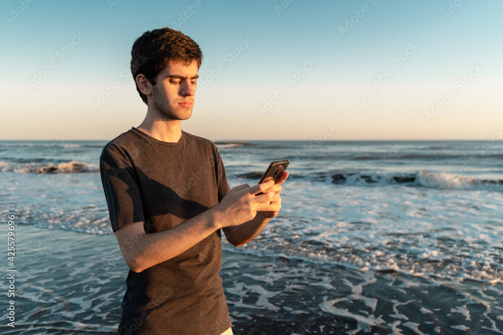 Young man standing on the seashore texting at sunset.