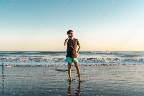 Young man standing on the seashore at sunset talking on the phone.