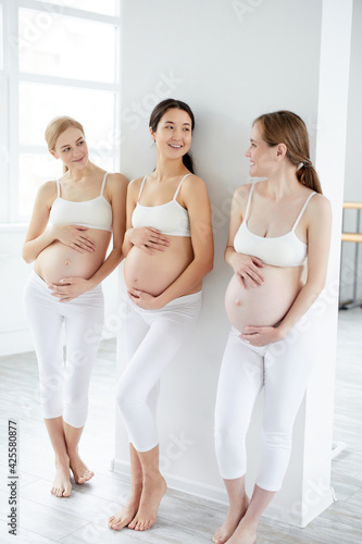 Portrait of three attractive young pregnant women standing in studio in white sportive outfit