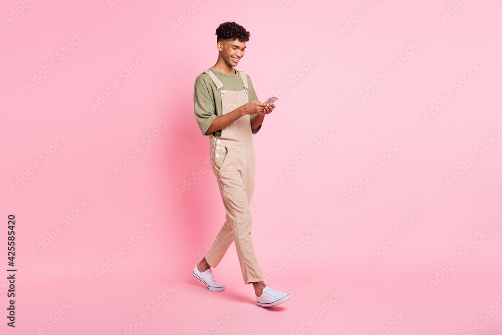 Full length body size photo of young man in overall smiling walking texting message on cellphone isolated on pastel pink color background Full length body size photo of young man in overall smiling walking texting message on cellphone isolated on pastel pink color background