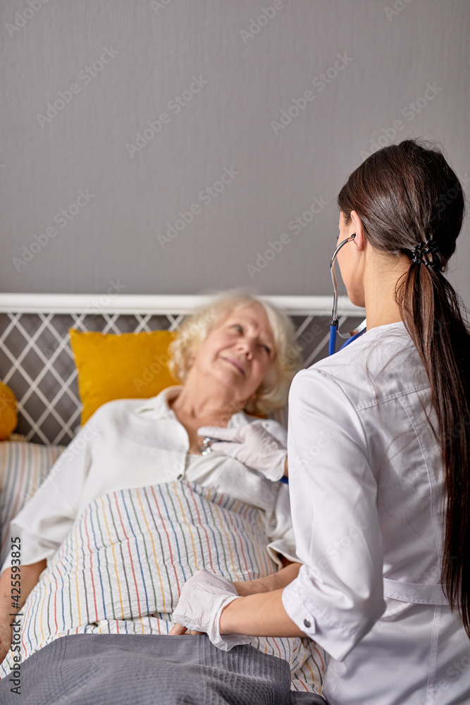 rear view on helpful female nurse checking sounds in heart and lungs of sick senior woman lying on bed rear view on helpful female nurse checking sounds in heart and lungs of sick senior woman lying on bed