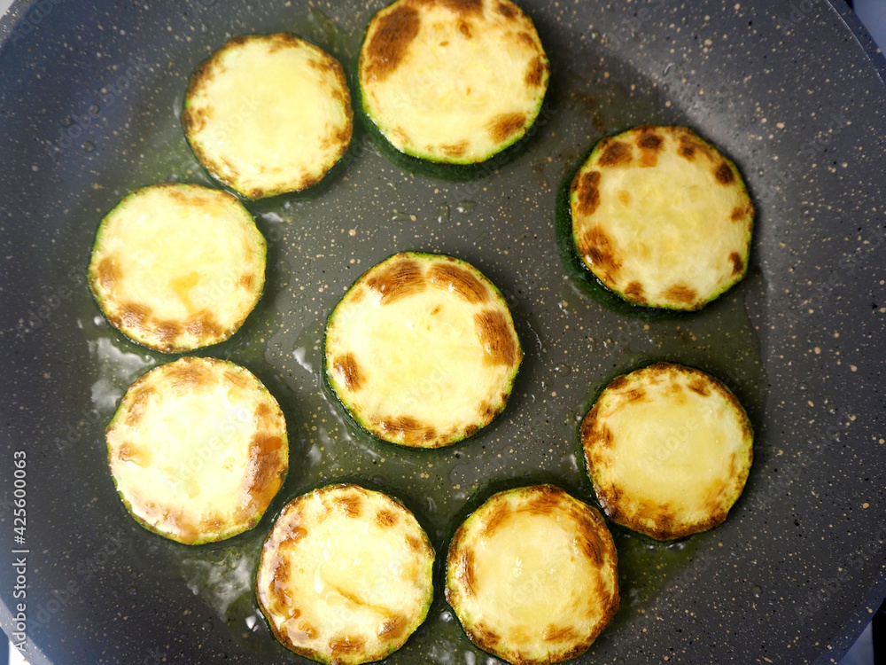the process of frying green sliced zucchini slices in vegetable oil in a gray frying pan top view . vegetarianism the process of frying green sliced zucchini slices in vegetable oil in a gray frying pan top view . vegetarianism