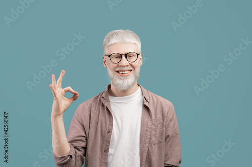 Everything is okay. Joyful albino guy doing approval ok gesture with fingers, showing that he is fine, blue studio background photo