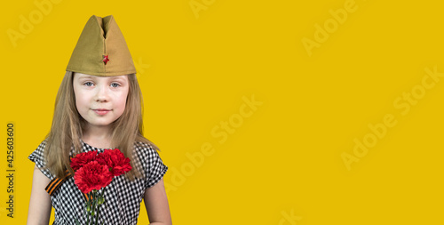 9th May. A little girl in a garrison cap with a St.George ribbon with red carnations stands on a yellow background. Victory Day. Memory of the Great Patriotic War. Soft Focus. Banner. copy space