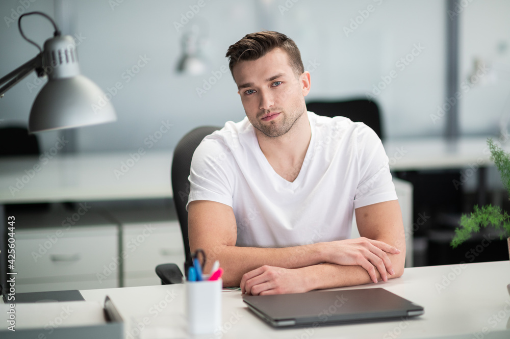 An attractive man in an office at a desk in a white t-shirt and with a laptop An attractive man in an office at a desk in a white t-shirt and with a laptop