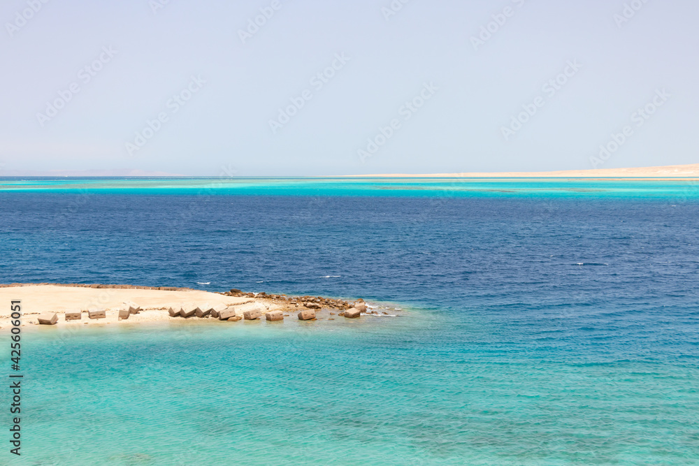 Seascape, panoramic view of the azure waters of the Red Sea, turquoise coral reefs in the distance, white desert sand on the horizon, part of the rocky coast in the foreground