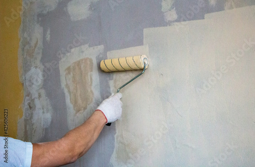 mans hand in cloth glove with paint roller applies fresh paint and primer to wall surface. process of applying water-based paint and primer with paint roller on surface for  interior design of room.