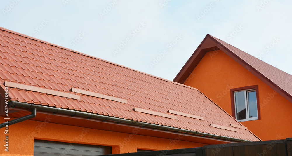 Metal roofing construction: A red metal gable roof with snow stoppers, snow guards and roof gutters of an orange painted house.