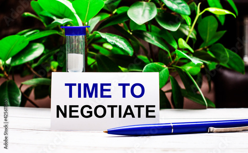 Against the background of green leaves of the plant, there is a pen on the table, an hourglass and a card with the inscription TIME TO NGOTIATE photo