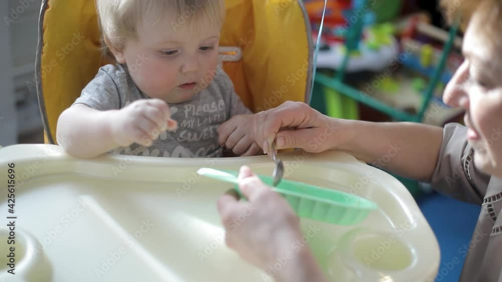 Grandmother lovingly feeds her grandson, the baby sits on a high chair. Close-up Grandmother lovingly feeds her grandson, the baby sits on a high chair. Close-up