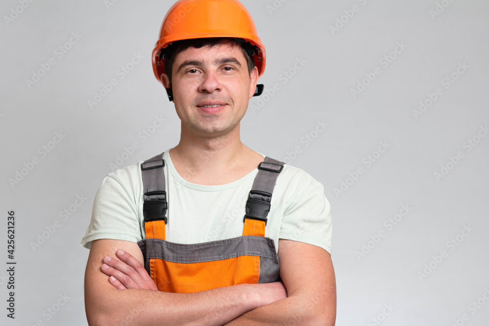confident young construction worker in hard hat and overalls with arms folded on the chest on grey studio background