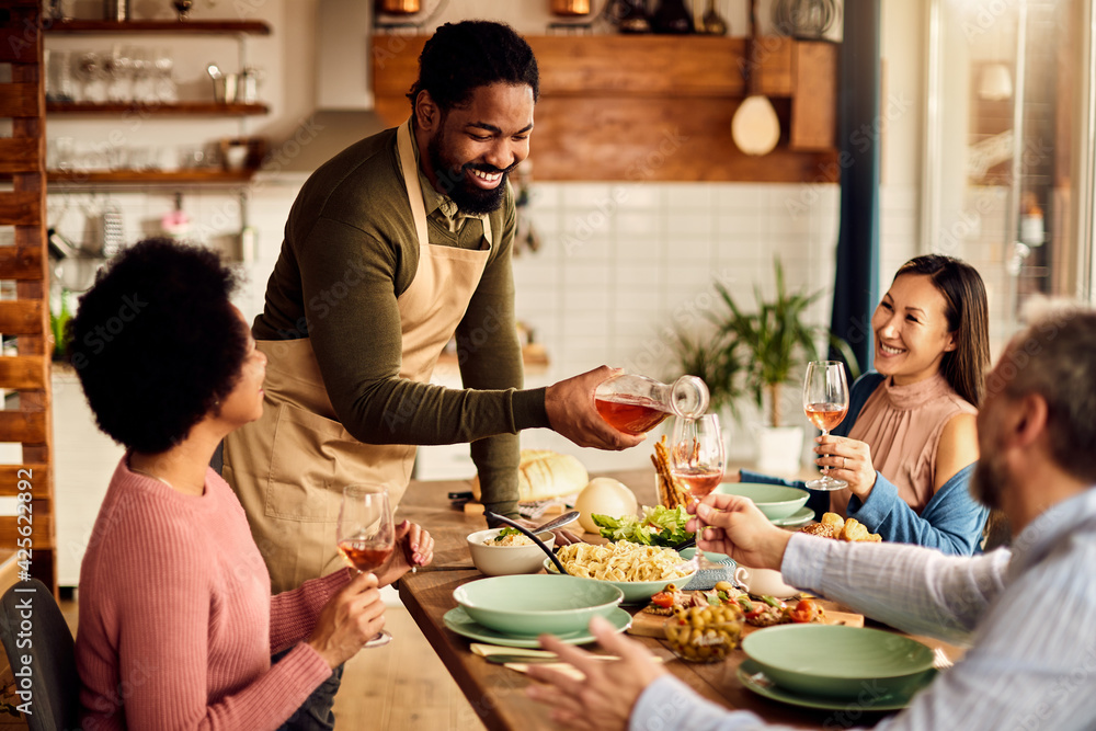 Happy African American man serving wine to his friends while having lunch at dining table.